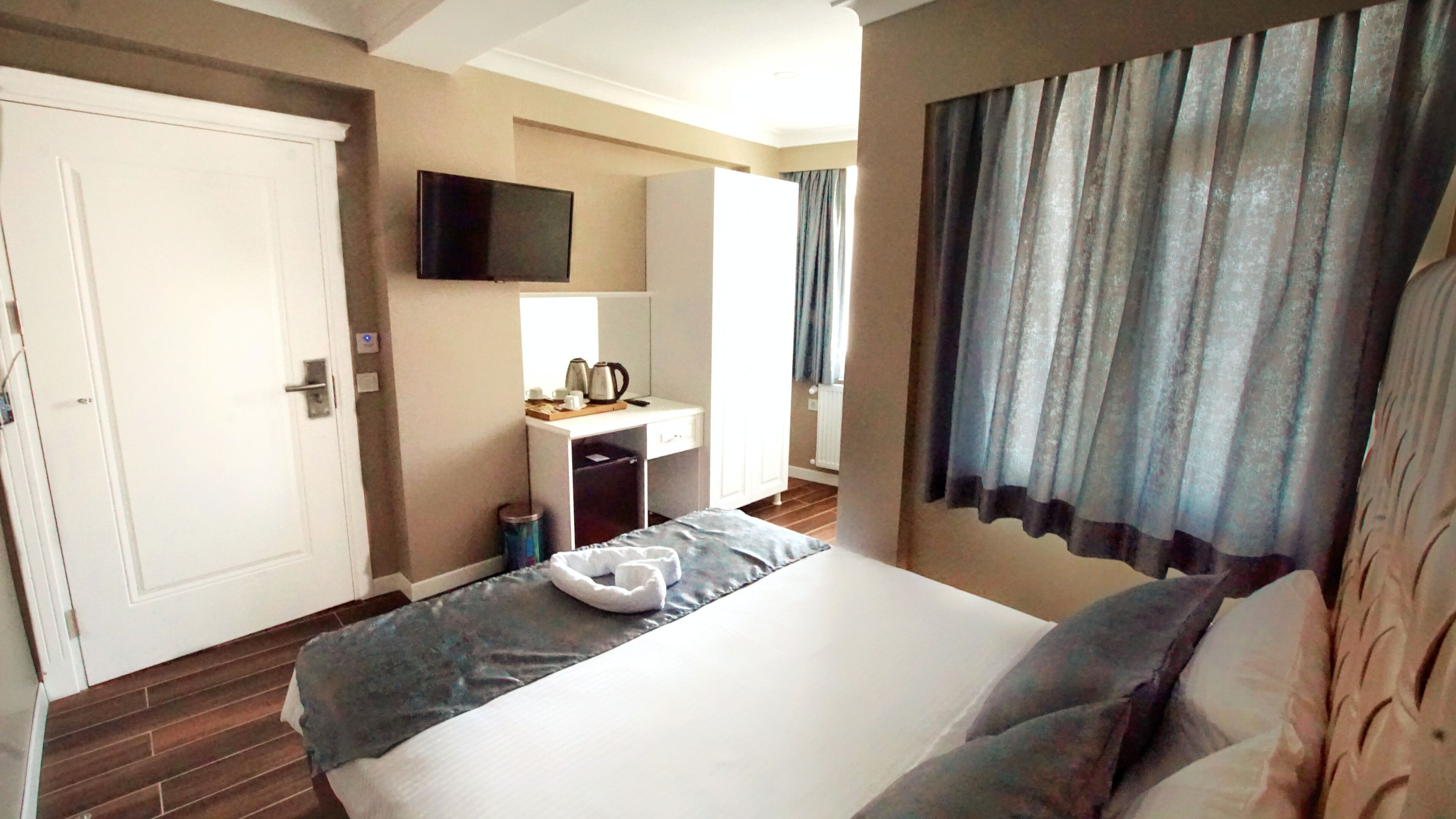 Une chambre d’hôtel avec un lit, un bureau avec une bouilloire, une télévision et une fenêtre avec des rideaux.