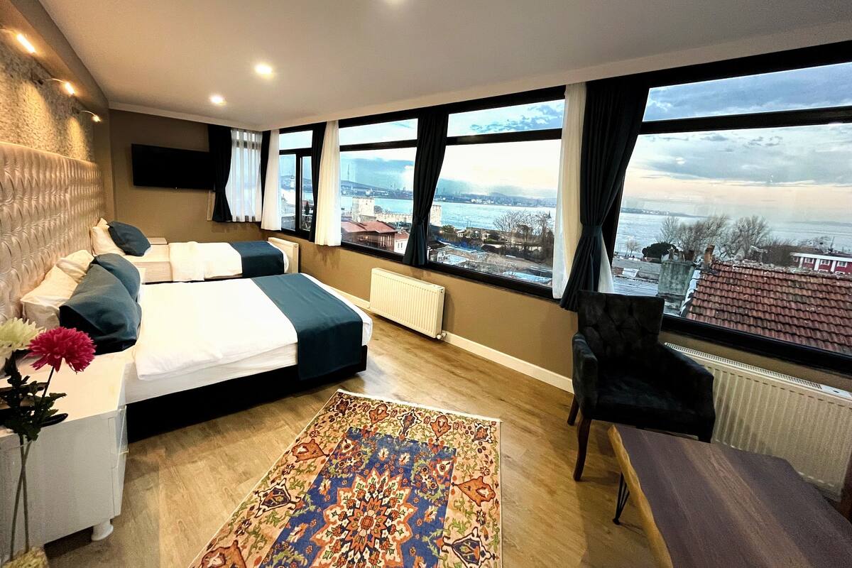 Quarto de hotel moderno com uma cama grande, vista para a cidade e uma janela ampla com cortinas.