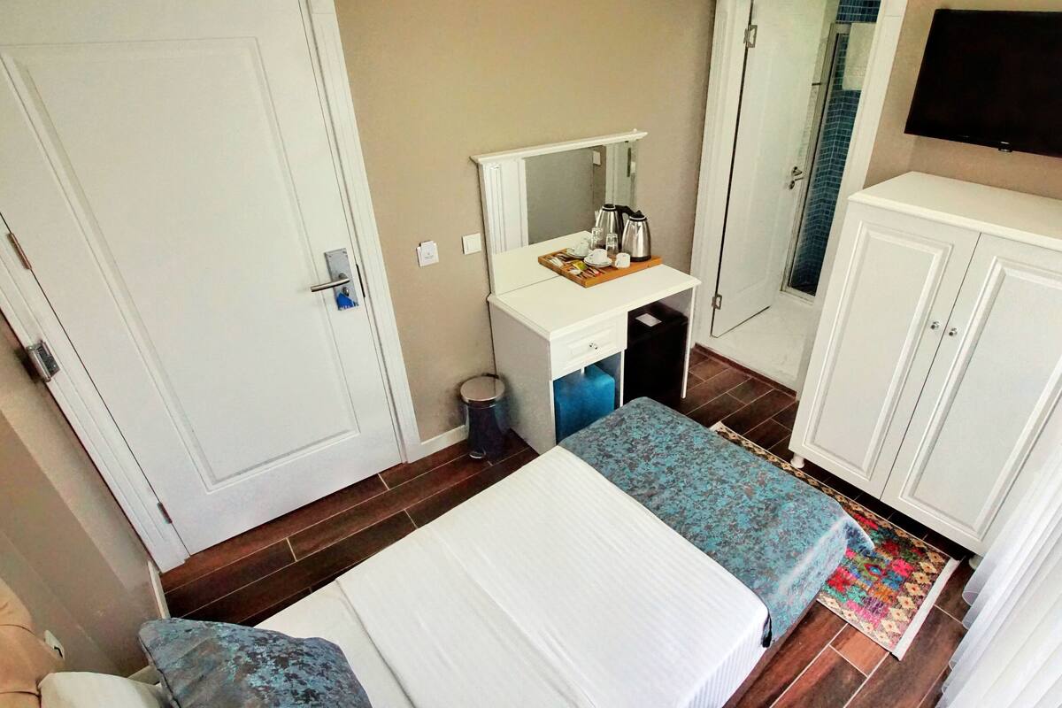 Quarto de hotel com cama, pequena escrivaninha com espelho, guarda-roupa e televisão na parede.