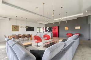 Una sala moderna con sofás grises, una mesa de centro de vidrio y sillas rojas. Se observa un comedor con sillas de madera y una mesa. En el fondo, se distingue una cocina con armarios rojos.