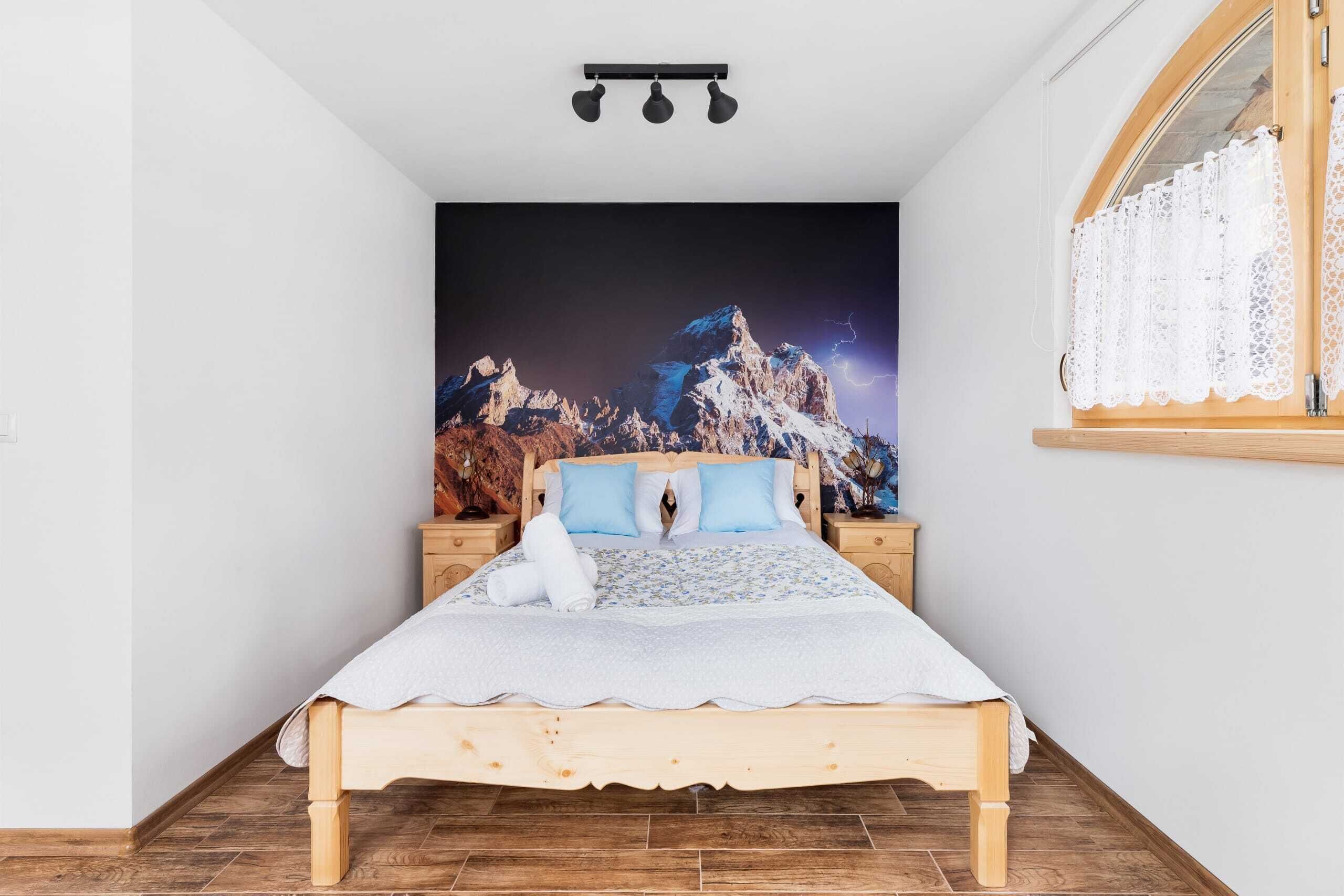 A bedroom with a wooden bed, a nightstand, a wall-mounted lamp, and a window with lace curtains.
