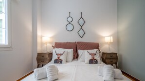 Ein ordentlich bezogenes Bett mit weißen Laken, zwei dekorative Kissen mit einem Hirschmuster, zwei Nachttische mit Lampen und Wandbilder über dem Bett.