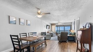 A spacious living room with a wooden dining table, chairs, and a ceiling fan. There are framed pictures on the wall, a large window with a view, and a sofa. - Edgewater 41 (Gulf Shores)