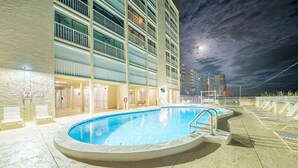 A swimming pool with a sunken area, surrounded by a building with multiple balconies and a view of the city skyline at night. - Edgewater 41 (Gulf Shores)