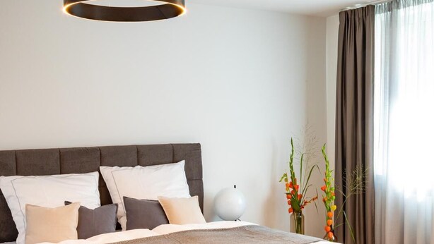 Een moderne slaapkamer met een groot bed, twee hanglampen, een nachtkastje en een vaas met bloemen.
