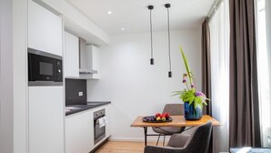 Una cocina moderna con armarios blancos, encimera negra y microondas integrado. Hay una mesa de comedor con un florero y un plato de fruta. El ambiente tiene suelo de madera y una ventana con cortinas.