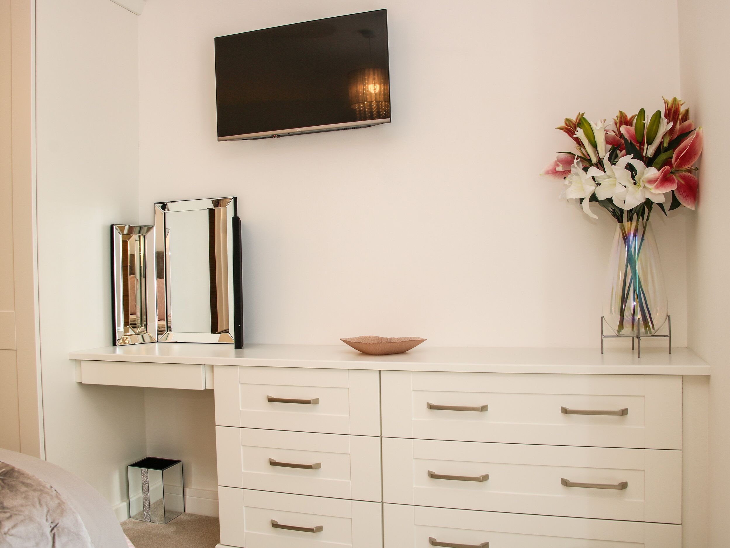Une commode blanche avec des tiroirs et un miroir, un téléviseur à écran plat fixé au mur et un vase avec des fleurs posé sur la commode.