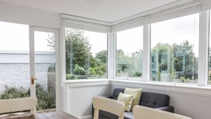 A sunroom with a view of a lake, a wooden table, and a grey sofa with cushions. - Castlebrae (Jedburgh)