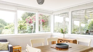 A bright room with a wooden dining table, white chairs, and a small wooden stool. Large windows offer a view of greenery and flowers outside. - Castlebrae (Jedburgh)