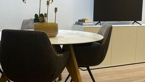A modern dining area with a round table, two chairs, a vase with white flowers, and a flat-screen TV on a stand.