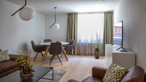 A modern living room with a brown leather sofa, a glass coffee table, and a dining area with a round table and chairs. There is a large window with curtains, a flat-screen TV on a stand, and a vase with plants on the coffee table.