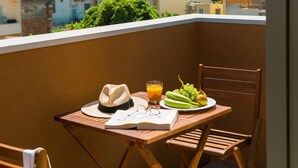A balcony with a table, a chair, a hat, a pair of glasses, a book, a glass of orange juice, and a plate of green grapes and a pear. - Thallos the Boutique (Chania)