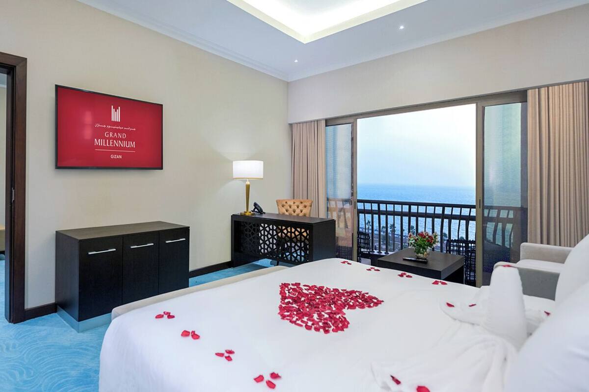 A hotel room with a bed, a TV, a desk, a chair, and a balcony with a view of the sea.