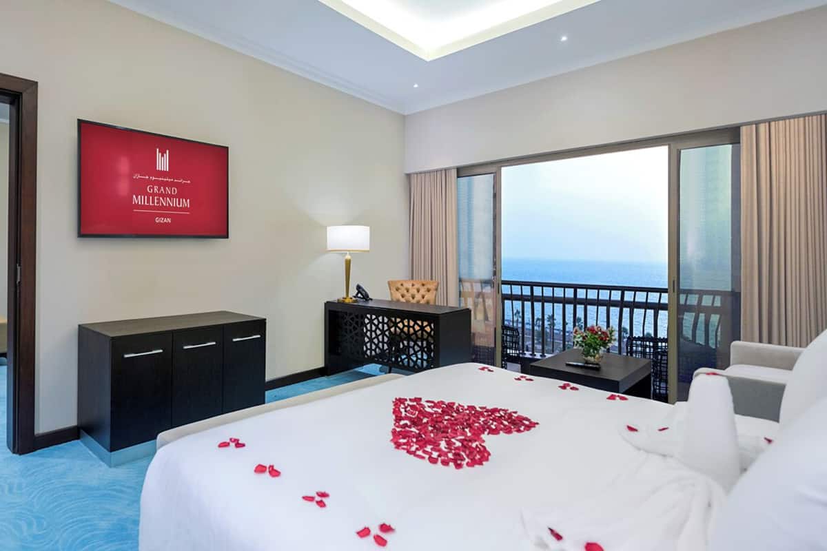A hotel room with a bed, a TV, a desk, a chair, and a balcony with a view of the sea.