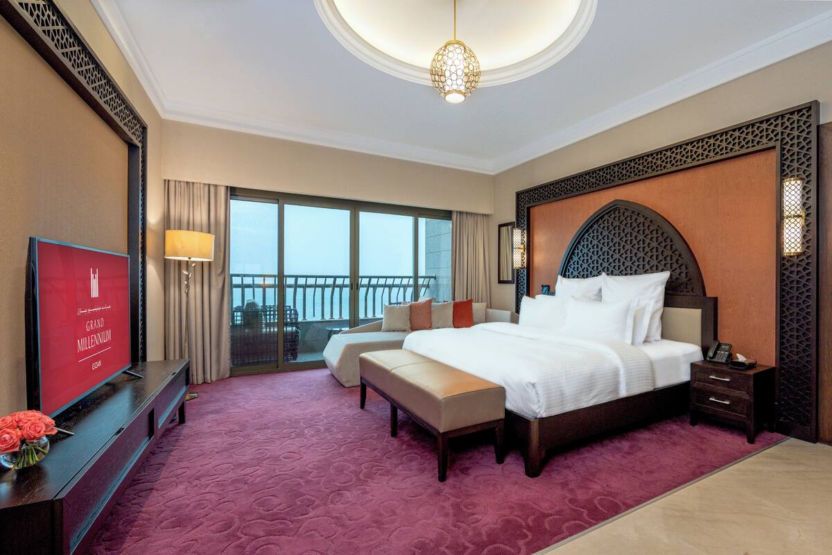 A hotel room with a large bed, a desk with a TV, a balcony with a view, and a decorative headboard.