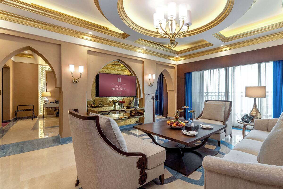 A luxurious hotel lobby with a marble floor, a central dining table with a fruit bowl, seating area with sofas, a bar, and a large TV screen displaying the hotel name.