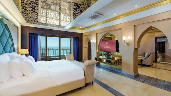 A hotel room with a large bed, a balcony with a view, a minibar, and a seating area with a TV.