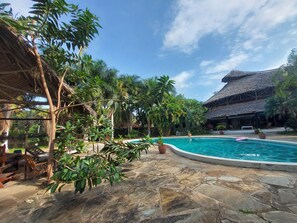 A pool area with stone flooring, surrounded by lush greenery and a thatched-roof building. - Red Lion (Malindi)