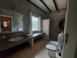 A bathroom with a large mirror, a sink, a toilet, and a window with blinds. - Red Lion (Malindi)