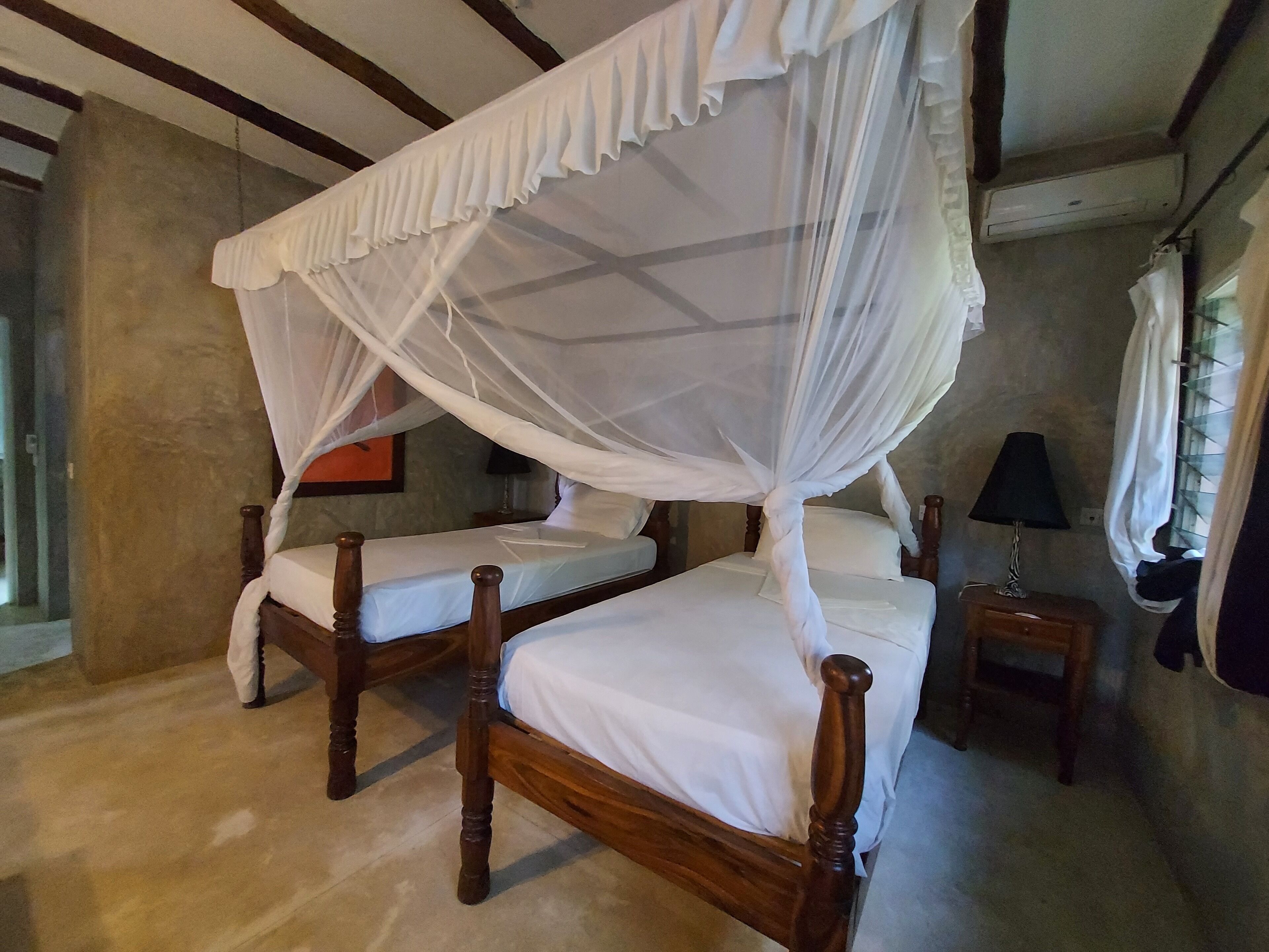A room with two wooden beds, each with a canopy. There is a small table with a lamp beside one bed.