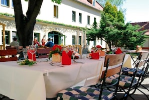 An outdoor dining area with tables set for multiple guests, surrounded by trees and a building facade. - Gutshof Hauber (Dresden)