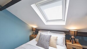 Ein Schlafzimmer mit Oberlicht, einem Bett mit gemusterten Textilien, zwei Nachttischen mit Lampen und einem Nachttisch aus Holz.