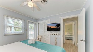 Un dormitorio con cama, ventilador de techo, televisor y vista a otra habitación.