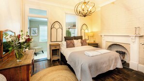 Une chambre à coucher comprenant un lit, des tables de chevet, un miroir, un vase avec des fleurs, une cheminée et un lustre.