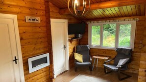 A cozy cabin interior with wooden walls, a fireplace, a television, and a window overlooking greenery. - Snowdonia Log Cabin-sleeps 4-garden-pets (Blaenau Ffestiniog)