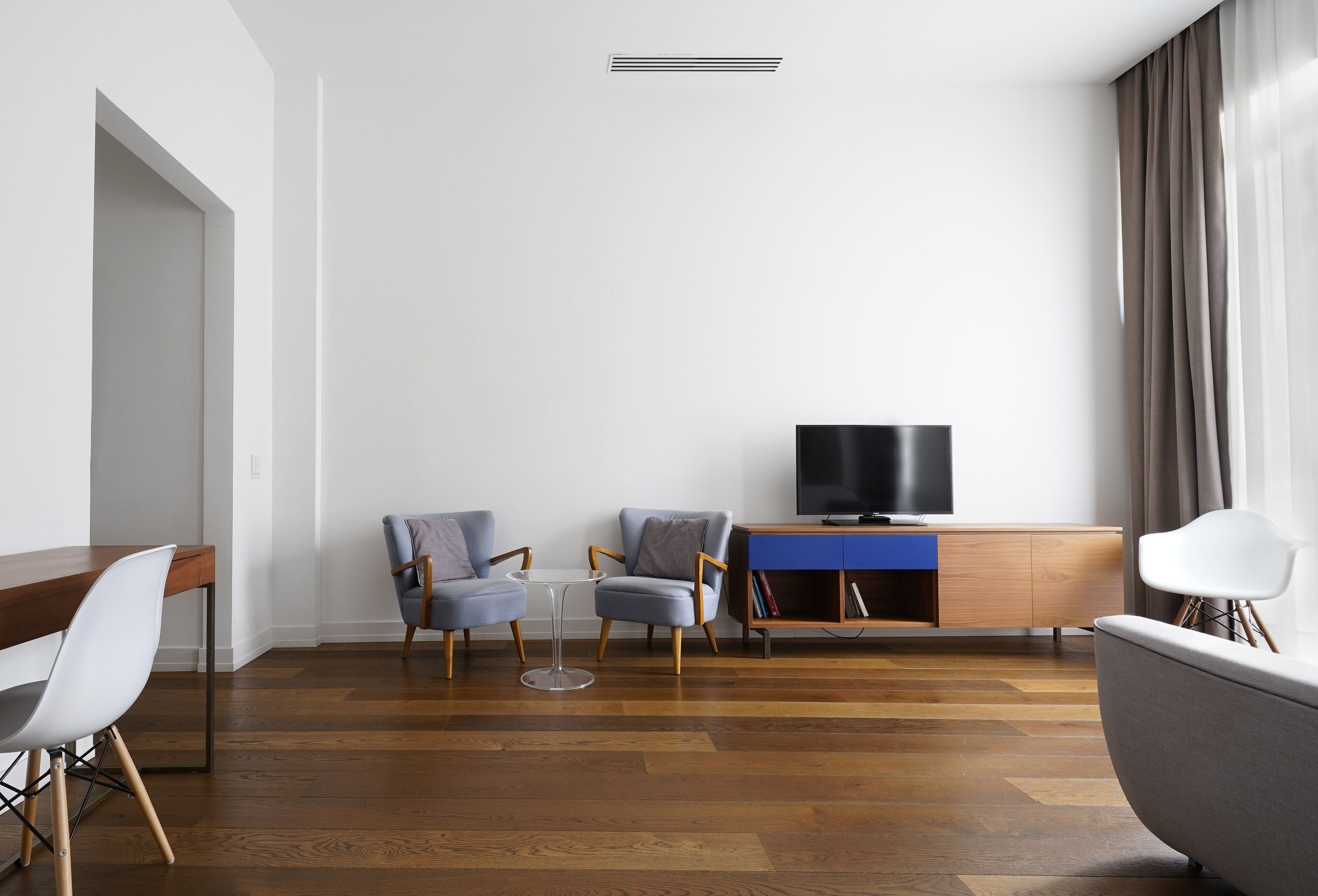 Una sala de estar moderna con piso de madera, una mesa de comedor con sillas, un televisor sobre un mueble de madera y un ventanal con cortinas.