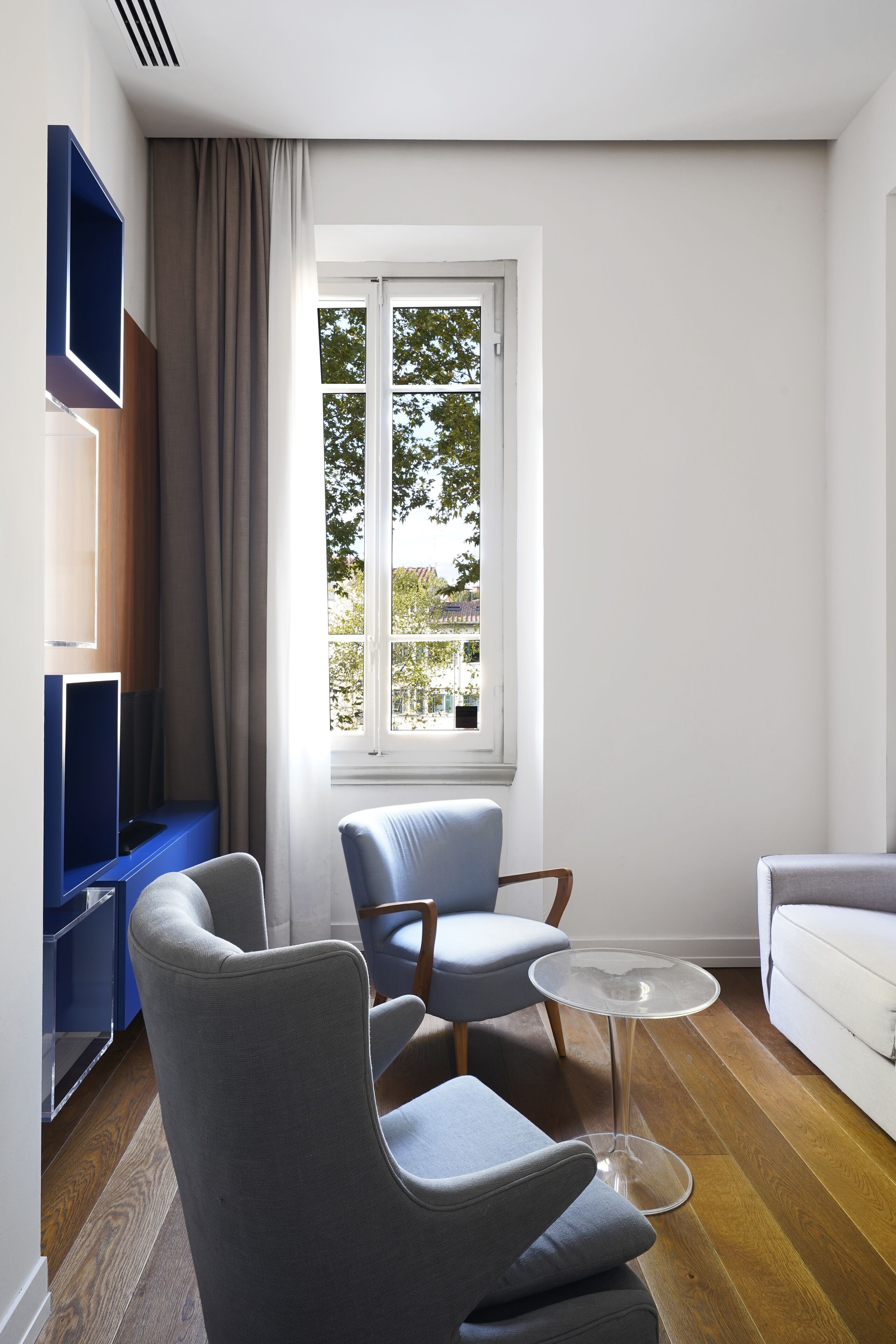 A modern living room with a large window, a grey sofa, a blue armchair, and a small glass table.