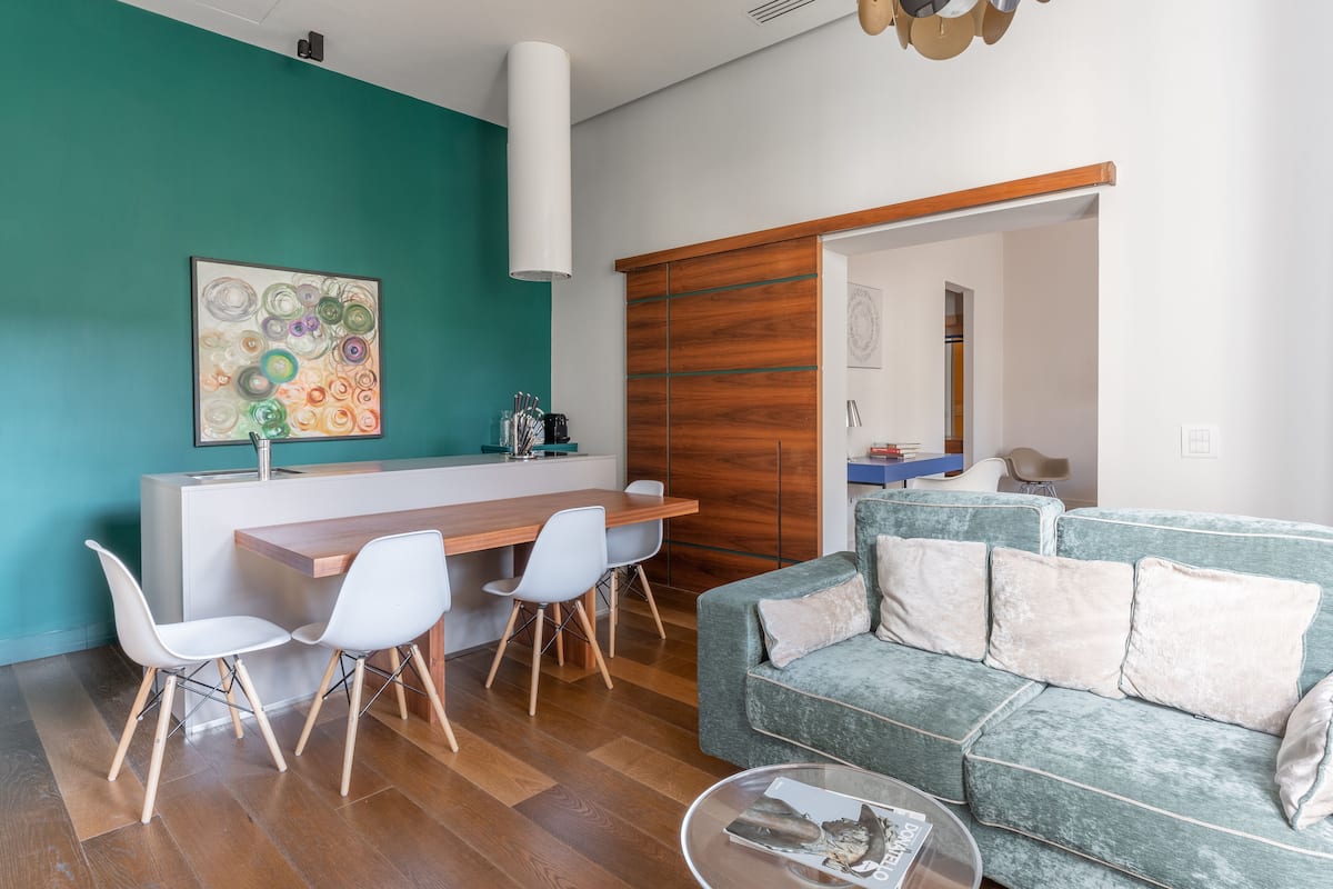 A modern living room with a teal accent wall, a wooden dining table with chairs, a green sofa with cushions, and a glass coffee table.