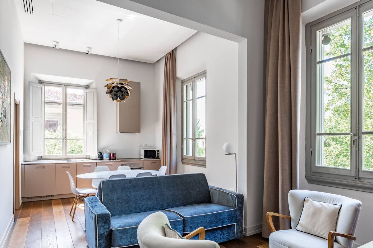 A modern living room with a blue sofa, white armchairs, and a wooden coffee table. There is a kitchen area with a dining table and chairs, and large windows providing natural light.
