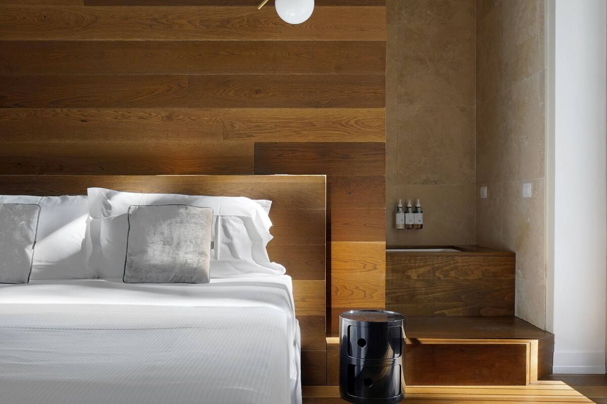 A modern bedroom with a wooden headboard, a bed with white linens, a bedside table, and a small seating area.