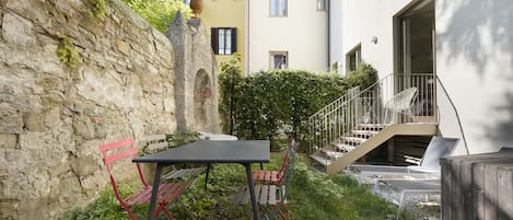 A courtyard with a stone wall, a table, and red chairs. There is a staircase leading to an upper level with balconies.