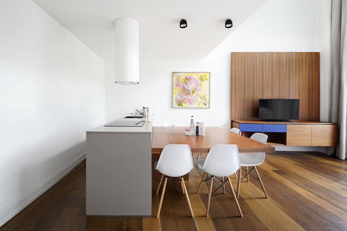 A modern kitchen with a central island, white chairs, a wooden dining table, and a built-in TV unit.