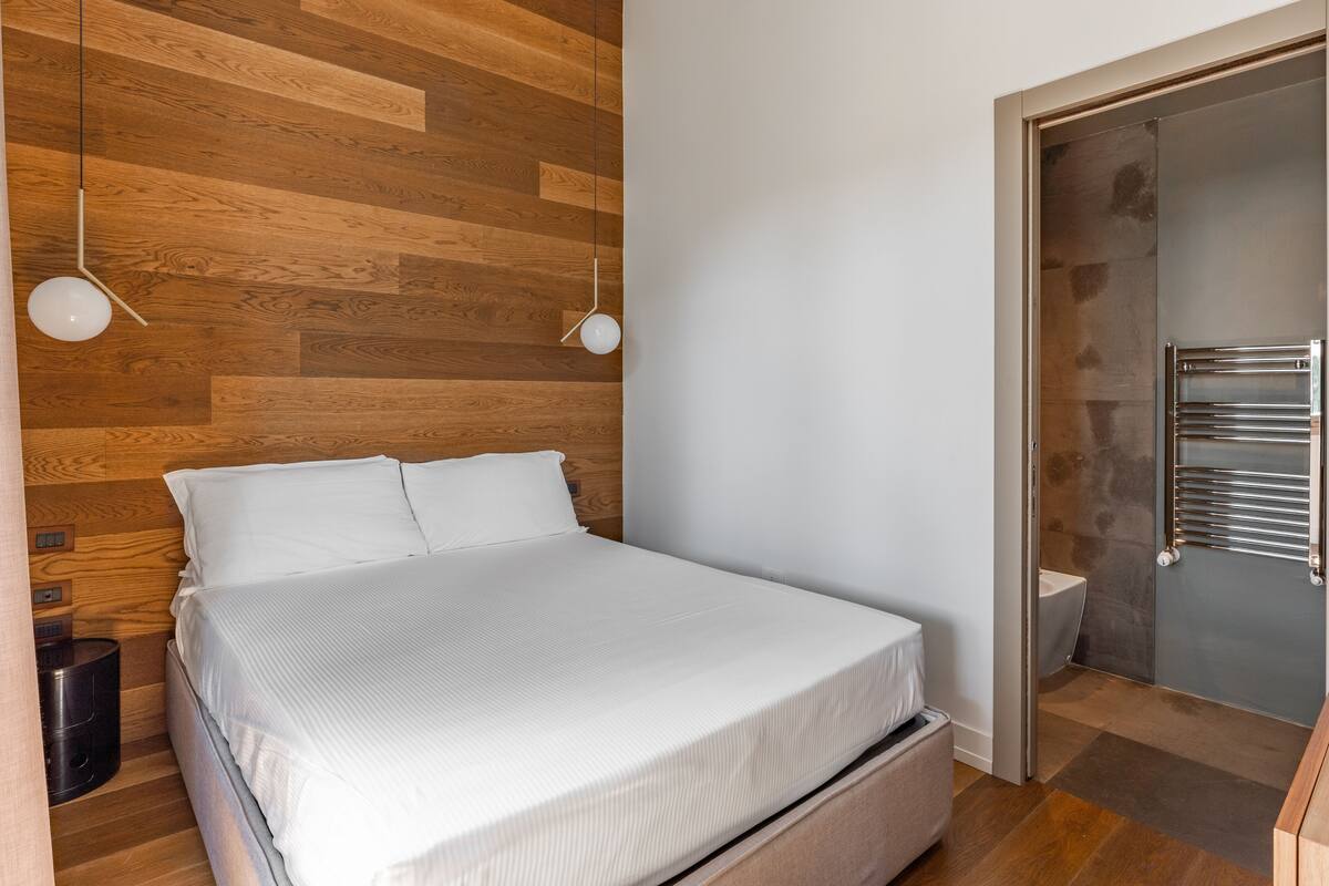 A modern bedroom with a wooden wall panel, a bed with white bedding, two bedside lamps, and a visible bathroom with a towel rack.