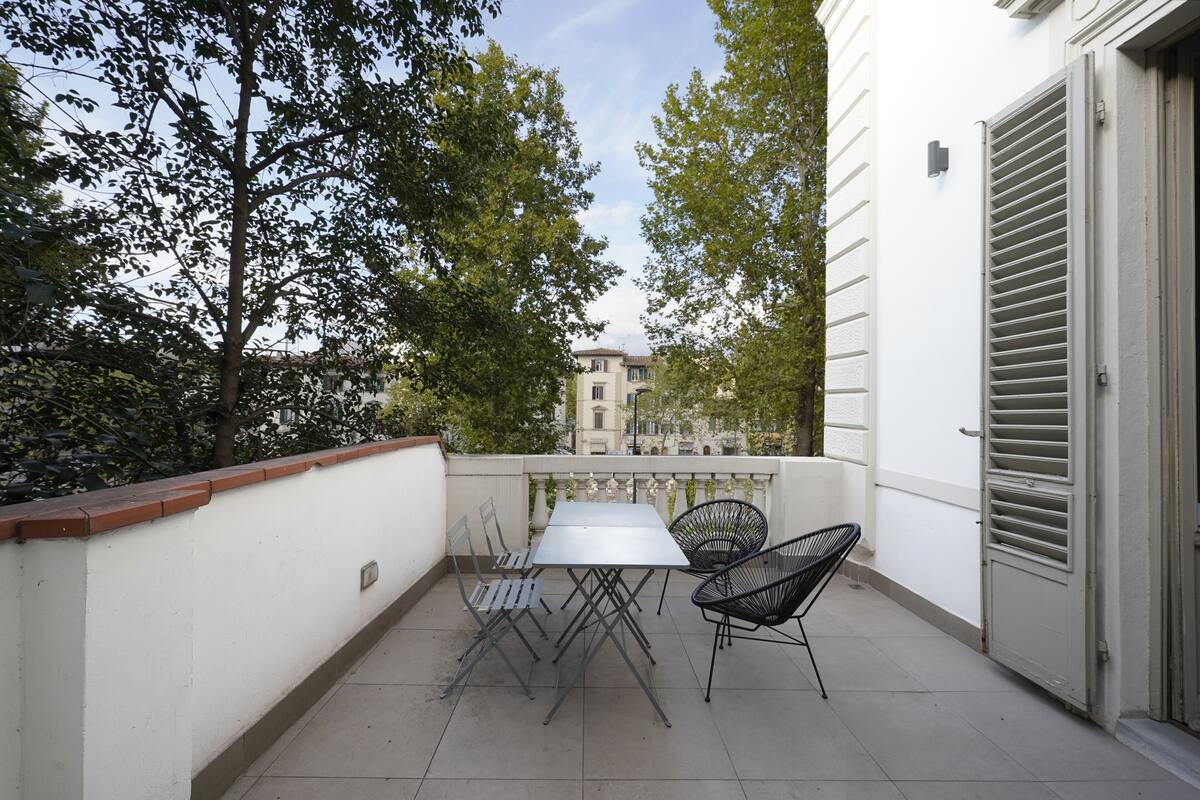 A balcony with a table and chairs, a white wall, and a door with shutters.