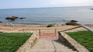 A stone staircase leading to a beach with a railing, a grassy area, and a clear view of the sea.