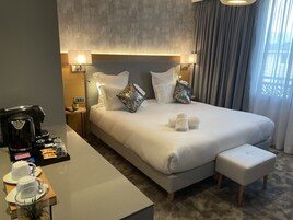 Una habitación de hotel moderna con una cama grande, una mesita de noche con cafetera y una pequeña mesa con tazas de té y aperitivos.