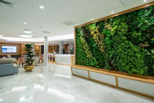 Un vestíbulo moderno con un jardín vertical grande y verde, un mostrador de recepción de mármol y un área de estar con sofás y sillones.