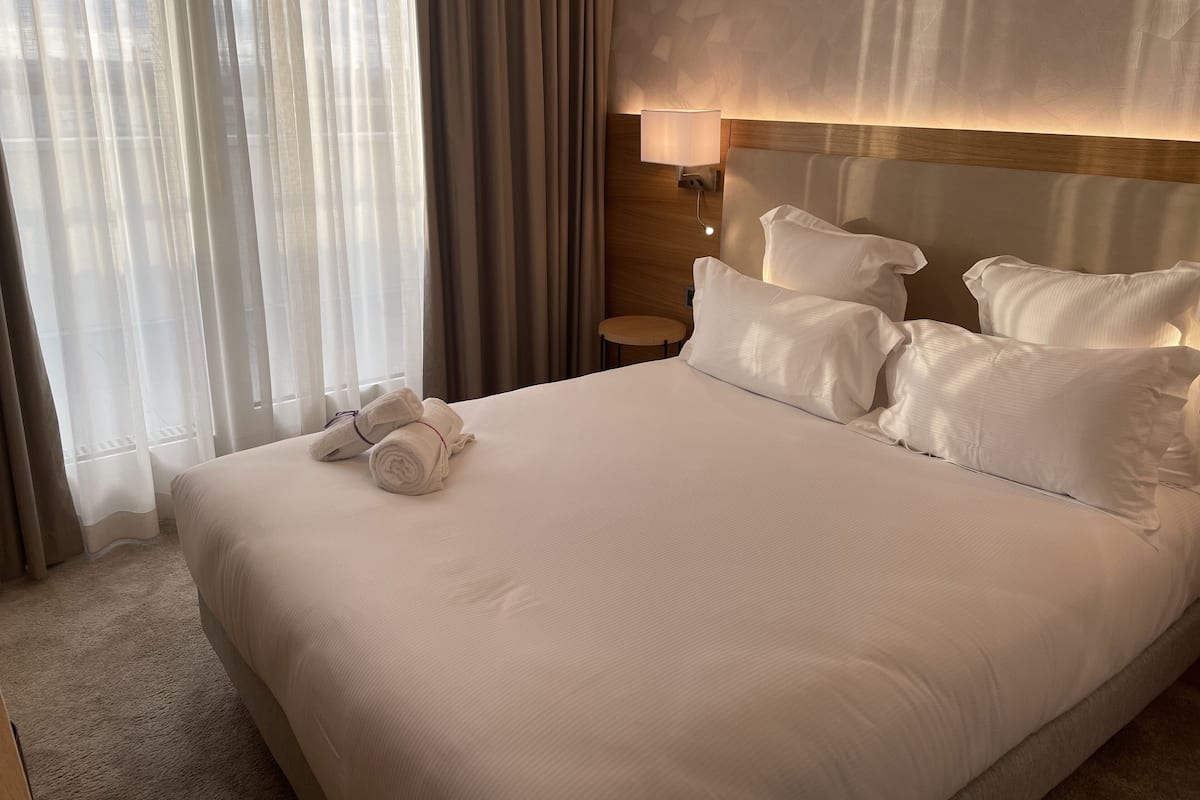 A neatly made bed with white linens and pillows, a bedside lamp, and a window with sheer curtains.