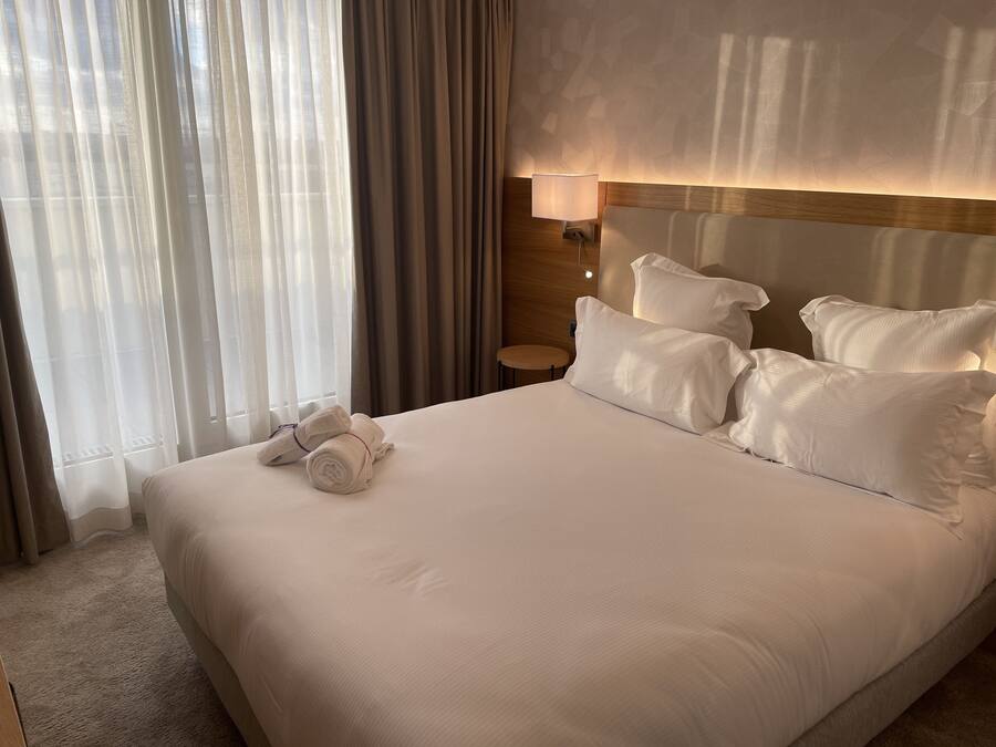 Un lit bien fait, avec du linge de lit blanc et des oreillers, une lampe de chevet et une fenêtre ornée de rideaux légers.