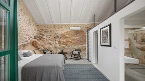 Une chambre avec un mur en pierre, un lit, un bureau, deux chaises et une échelle.