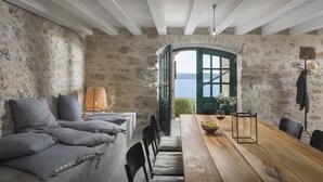 A rustic room with stone walls, a wooden dining table, a sofa with cushions, a lamp, and a view of the sea through a green door.
