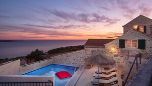 A stone villa with a swimming pool, a thatched umbrella, and a view of the sea at sunset.