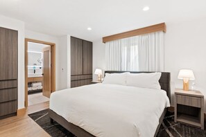Une chambre d’hôtel moderne avec un grand lit, des tables de chevet avec des lampes, une armoire en bois et une salle de bain visible à travers une porte ouverte.