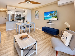 A modern living room with a wooden floor, a dining area, a kitchen, and a TV displaying a tropical beach scene.