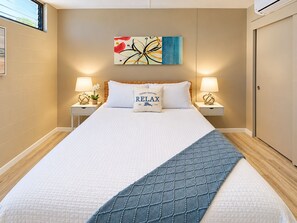 A neatly made bed with a white and blue quilt, two bedside lamps, a wall art piece, and a door on the right. - Airy Kihei Renovated Condo Ocean Views Spacious Lanai Near Kamaole Beach (Kihei)