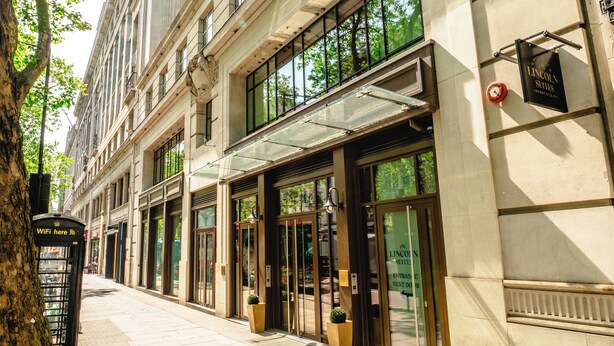 A multi-story building with large windows, a tree-lined sidewalk, and a sign indicating a Lincoln Square location. - The Lincoln Suites (London)